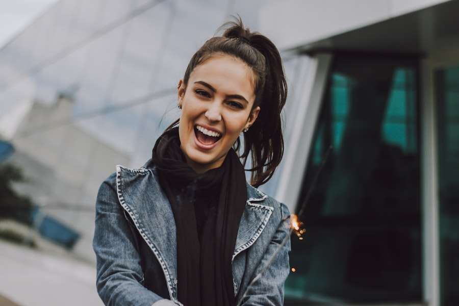 Suchmaschinenoptimierung Hannover - Eine Frau mit hohem Pferdeschwanz steht draußen und lächelt breit. Sie trägt eine Jeansjacke und einen schwarzen Schal. Im Hintergrund ist das moderne Glasgebäude von Webdesign Hannover zu sehen. Sie scheint eine Wunderkerze in der Hand zu halten, aus der kleine Funken sprühen. Local SEO Agentur Hannover.