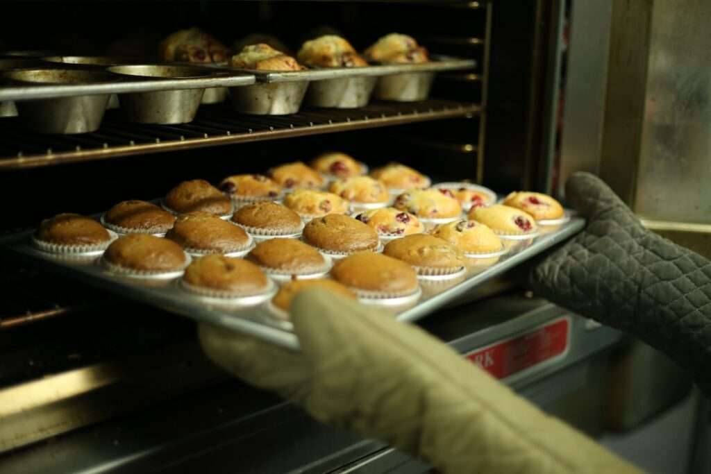 Suchmaschinenoptimierung Hannover - Eine Person mit Ofenhandschuhen hält ein Blech mit frisch gebackenen Muffins und stellt es in einen Ofen oder nimmt es aus dem Ofen. Die Ofentür ist geöffnet und im Ofen befinden sich weitere Muffins auf einem Blech. Local SEO Agentur Hannover.
