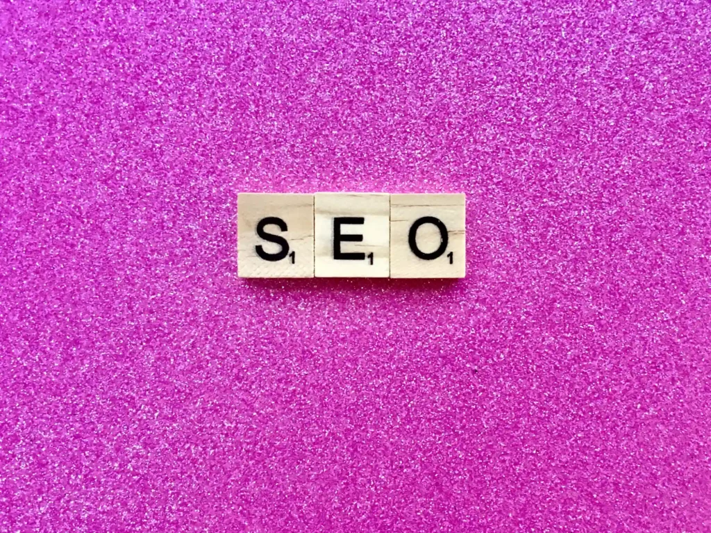 Suchmaschinenoptimierung Hannover - Drei Scrabble-Steine aus Holz mit dem Wort „SEO“ liegen auf einem rosa, glitzernden Hintergrund. In der unteren rechten Ecke jedes Steins ist ein Buchstabe mit einem Punktwert abgebildet. Local SEO Agentur Hannover.
