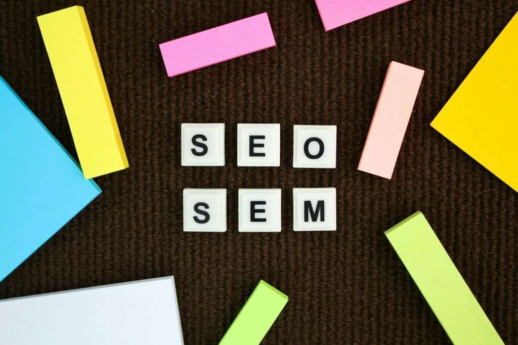 SEO Hannover - Die Kacheln mit den Buchstaben „SEO“ und „SEM“ sind auf einer braun strukturierten Oberfläche angeordnet und von bunten rechteckigen Papieren in Gelb, Rosa, Blau und Grün umgeben. Local SEO Agentur Hannover.