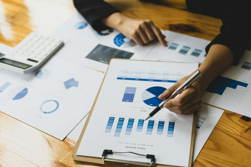 SEO Hannover - Eine Person mit einem Stift überprüft Diagramme und Grafiken auf einem Klemmbrett mit Verkaufsberichten und analysiert die Daten sorgfältig, um das Google-Ranking zu optimieren. Verschiedene blau getönte Grafiken sind über den Holztisch verteilt, und in der oberen linken Ecke ist ein Taschenrechner zu sehen. Local SEO Agentur Hannover.