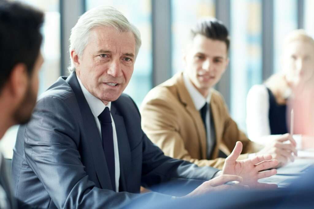 SEO Hannover - Eine Gruppe von vier Personen in formeller Kleidung sitzt um einen Tisch in einem Konferenzraum mit großen Fenstern. Ein älterer Mann mit grauem Haar spricht und gestikuliert mit einer Hand, während die anderen aufmerksam zuhören und möglicherweise SEO-Strategien für Anwälte diskutieren. Local SEO Agentur Hannover.