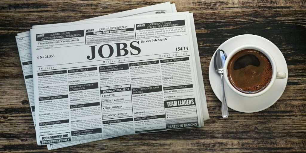SEO Hannover - Auf einer Holzoberfläche liegt ein Stapel Zeitungen, aufgeschlagen auf der Seite mit den Stellenausschreibungen. Die Seiten sind mit verschiedenen Stellenanzeigen gefüllt, die an Suchergebnisse von Google for Jobs erinnern. Neben den Zeitungen steht eine Tasse Kaffee mit einem Löffel auf der Untertasse. Local SEO Agentur Hannover.