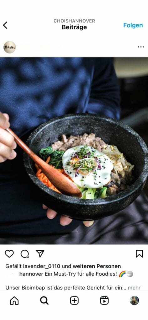 Webdesign Hannover - Eine Person hält eine mit Bibimbap gefüllte schwarze Steinschüssel, auf der Reis, geschnittenes Gemüse, Rindfleisch und ein Spiegelei liegen. In ein schwarzes Langarmshirt gekleidet und mit einem Holzlöffel in der Hand ist sie bereit für das perfekte Instagram-Feed-Foto. Der Hintergrund ist kunstvoll verschwommen. Local SEO Agentur Hannover.