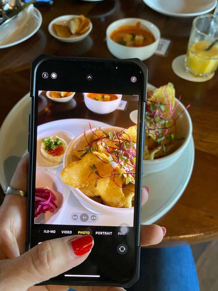 Webdesign Hannover - Eine Person mit einem Smartphone macht ein Foto von einem Teller voller knuspriger Chips, garniert mit Microgreens, Soße und eingelegtem Gemüse – perfekter Inhalt für Social Media Marketing. Im Hintergrund verleihen eine Schüssel Suppe und ein Getränk dem Holztisch Tiefe. Local SEO Agentur Hannover.