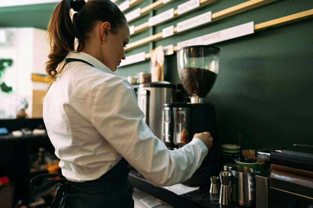 SEO Hannover - Ein Barista in weißem Hemd und Schürze bedient gekonnt eine Kaffeemühle hinter einer Café-Theke. Um ihn herum stehen verschiedene Werkzeuge zur Kaffeezubereitung sowie Regale mit beschrifteten Artikeln – jedes Detail wurde sorgfältig ausgewählt, um optimale SEO für Bilder zu gewährleisten. Local SEO Agentur Hannover.