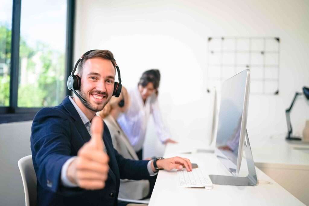 Webdesign Hannover - Eine Person aus dem Kontakt-Team sitzt mit Headset und Anzug an einem Computertisch, lächelt und streckt den Daumen nach oben. Zwei Kollegen arbeiten im Hintergrund, einer an einem anderen Schreibtisch. Große Fenster sorgen für natürliches Licht in diesem lebendigen SEO-Büroraum. Local SEO Agentur Hannover.