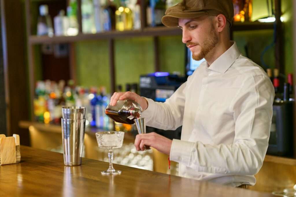 SEO Hannover - Ein Barkeeper in weißem Hemd und Mütze gießt fachmännisch Flüssigkeit aus einer Karaffe in ein Glas an der Holztheke. Die Szene, perfekt für SEO für Bilder-Enthusiasten, zeigt verschiedene Flaschen, die im Regal im Hintergrund aufgereiht sind. Local SEO Agentur Hannover.