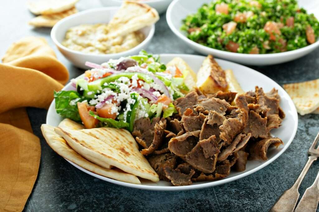 SEO Hannover - Ein Teller mit Gyros-Scheiben, einem frischen Salat mit Feta, Pita-Brot und einigen Kartoffelspalten steht verlockend da. Im Hintergrund begleiten Schüsseln mit Hummus und Taboulé neben einer gelben Serviette das Festmahl. Perfekt, um Küchendetails in SEO-Strategien für Bilder festzuhalten. Local SEO Agentur Hannover.