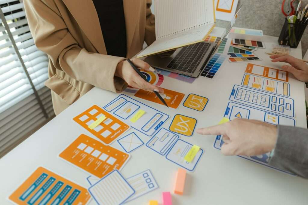 Webdesign Hannover - Zwei Personen sind an einem Schreibtisch voller bunter Papierausschnitte, Haftnotizen und verschiedener Designvorlagen vertieft in ein kreatives Designprojekt. Einer hält einen Laptop und ein Notizbuch, während der andere mit einem Stift darauf zeigt und laut grübelt: „Was genau ist eine Vorlage?“ Local SEO Agentur Hannover.