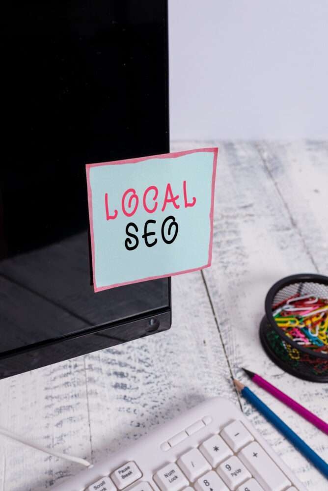 Webdesign Hannover - An der Ecke eines Computerbildschirms klebt ein Notizzettel mit dem Wort „Local SEO“. Darunter ist eine weiße Tastatur zu sehen. Rechts steht ein Behälter mit bunten Büroklammern und zwei Bleistiften auf einer weißen Holzoberfläche. Local SEO Agentur Hannover.