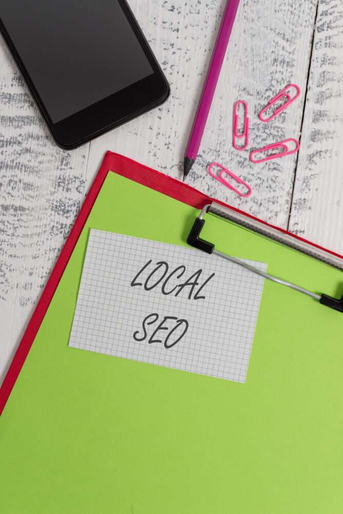 Webdesign Hannover - Ein Smartphone, ein rosa Bleistift und rosa Büroklammern liegen auf einer weißen Holzoberfläche. Auf einem roten Klemmbrett mit einem grünen Blatt liegt eine Notiz mit der Aufschrift „LOCAL SEO“. Local SEO Agentur Hannover.