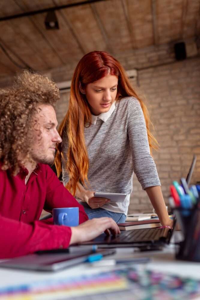 Webdesign Hannover - Ein Mann mit lockigem Haar in einem roten Hemd arbeitet an einem Laptop an einem Schreibtisch mit einer blauen Tasse und konzentriert sich auf die Neugestaltung einer Website. Eine Frau mit langen roten Haaren in einem grauen Pullover blickt auf seinen Bildschirm. Sie befinden sich in einem Raum mit einer Ziegelwand und einer Holzdecke, umgeben von verschiedenen Büromaterialien. Local SEO Agentur Hannover.