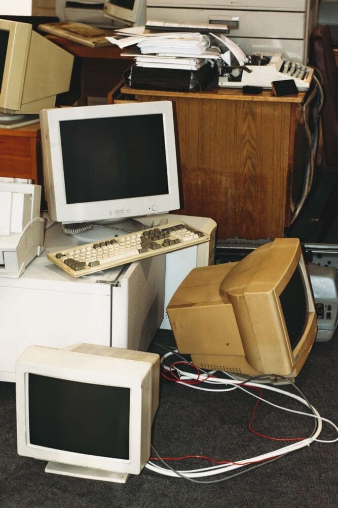 Webdesign Hannover - Ein überfüllter Raum mit mehreren alten CRT-Monitoren und Tastaturen, die auf einem Drucker gestapelt sind, erinnert an eine vergangene Ära vor dem Relaunch der Website. Einige Monitore sind eingewickelt, Kabel schlängeln sich über den Boden. Im Hintergrund ist ein Holzschreibtisch unter Stapeln von Papieren und weiteren elektronischen Geräten für das nächste Projekt der Agentur begraben. Local SEO Agentur Hannover.