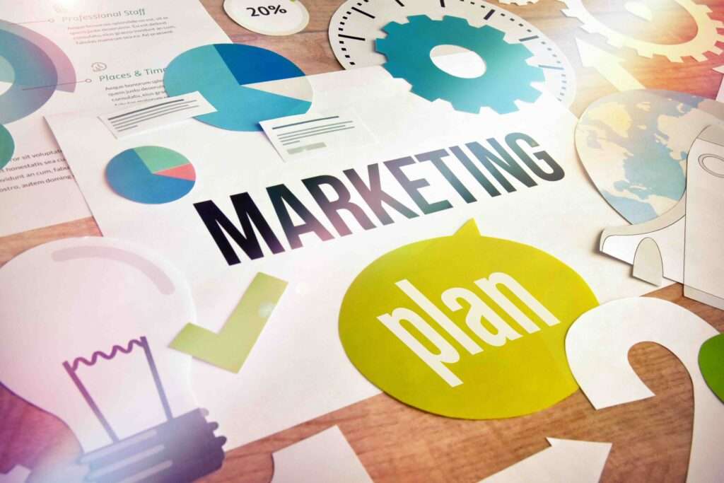 Webdesign Hannover - Eine Collage aus marketingbezogenen Elementen auf einer Holzoberfläche, mit den Worten „MARKETING“ in fetten schwarzen Buchstaben und „Plan“ in einer grünen Sprechblase. In der Nähe umgeben Symbole wie eine Glühbirne, ein Zahnrad und ein Globus die Szene – eine perfekte Inspiration für jede Marketingfirma in der Nähe. Local SEO Agentur Hannover.