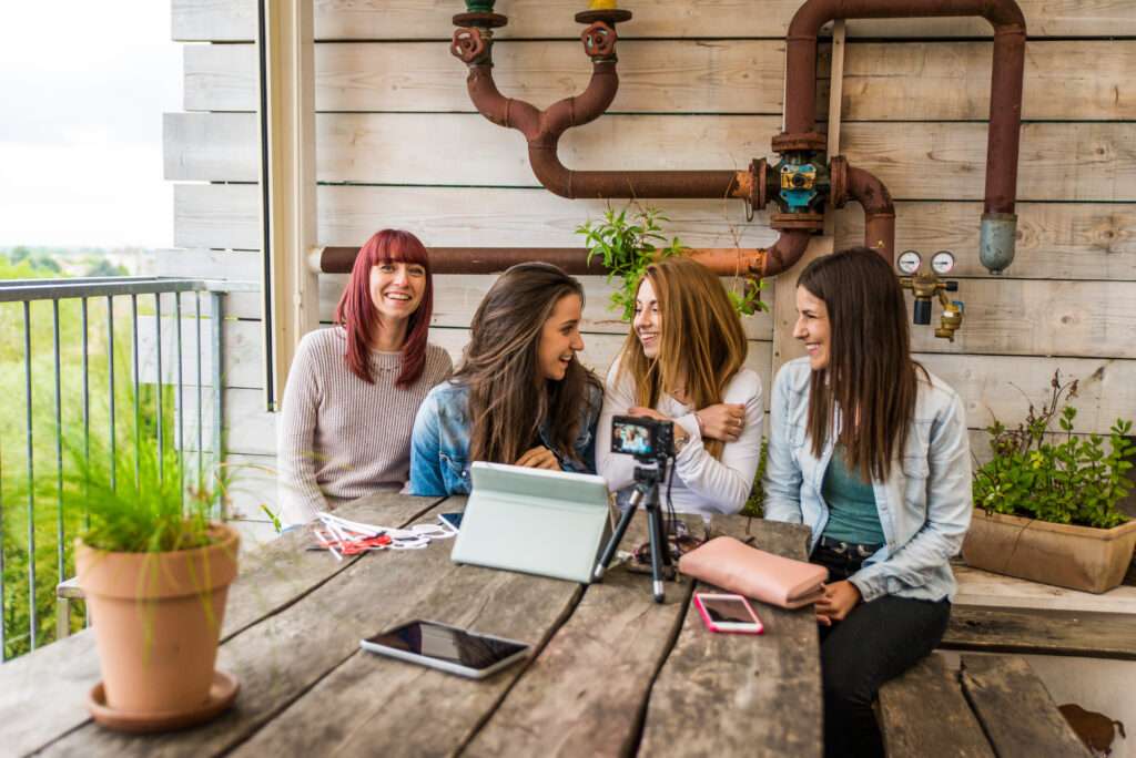 Webdesign Hannover - Vier Personen sitzen an einem Holztisch auf einem Balkon und unterhalten sich angeregt. Auf dem Tisch liegen eine Kamera auf einem Stativ und Tablets, die scheinbar über Website-Analysen diskutieren, während Topfpflanzen und freiliegende Rohre der Umgebung Charme verleihen. Local SEO Agentur Hannover.