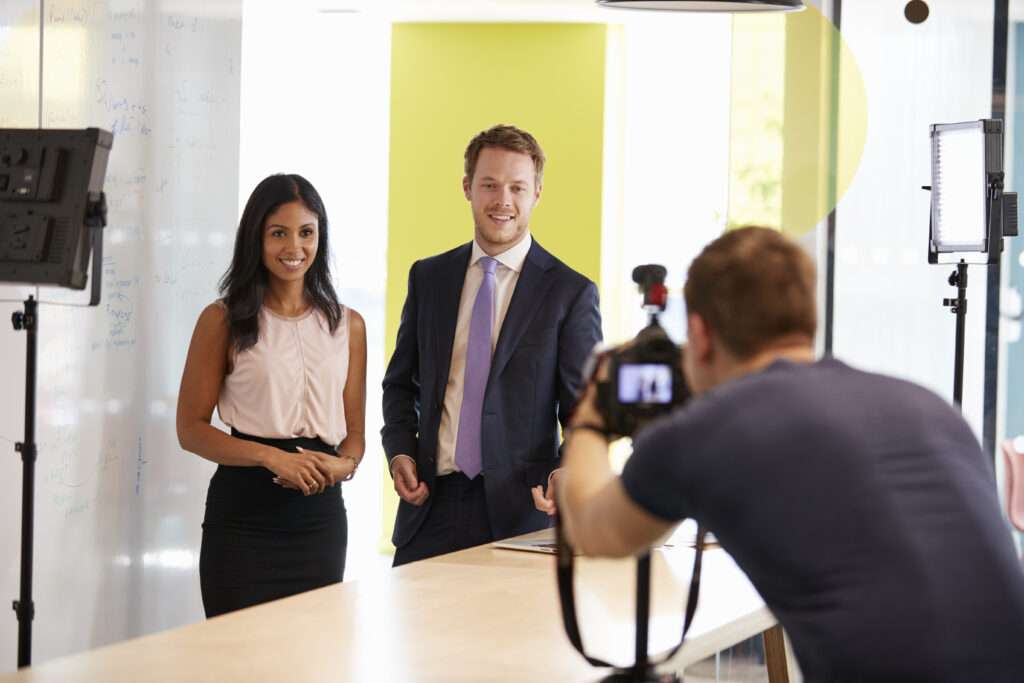Webdesign Hannover - Zwei Personen stehen nebeneinander in einem modernen Büro. Die eine trägt Bluse und Rock, die andere Anzug und Krawatte. Sie lächeln einem Business Fotograf Hannover zu, der ihren Moment einfängt. Im Hintergrund sind helle Lichter und ein Whiteboard zu sehen. Local SEO Agentur Hannover.